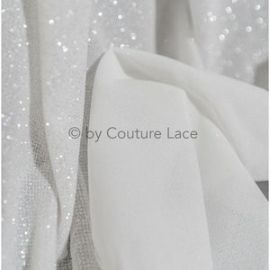 May include: White sheer fabric with a sparkly, glittery texture. The fabric is folded and draped, showing the shimmery effect of the glitter.