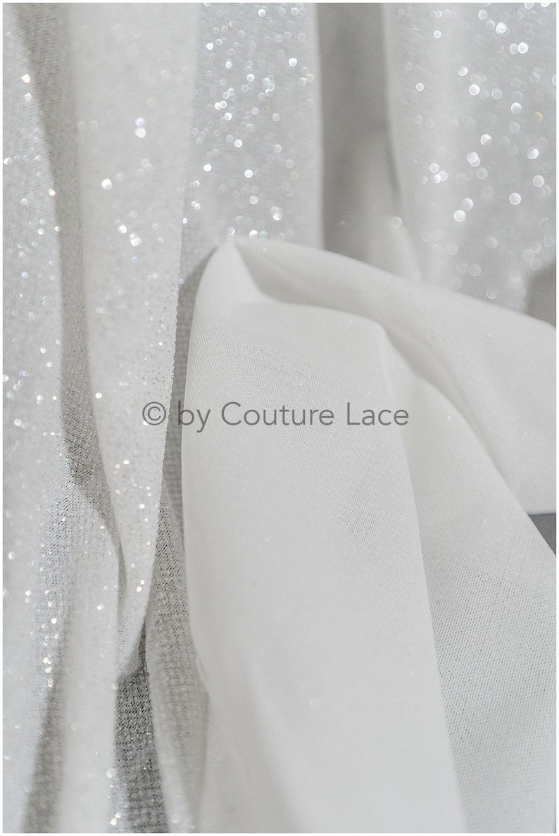 May include: White sheer fabric with a sparkly, glittery texture. The fabric is folded and draped, showing the shimmery effect of the glitter.