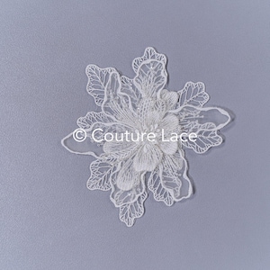 May include: White lace applique with a floral design. The applique features a large flower in the centre surrounded by smaller leaves. The applique is perfect for adding a touch of elegance to any project.