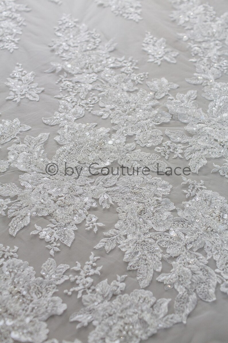 May include: White beaded lace fabric with a floral pattern. The lace is delicate and intricate, with small beads and sequins scattered throughout. The fabric is perfect for wedding dresses, bridal accessories, and other special occasion garments.