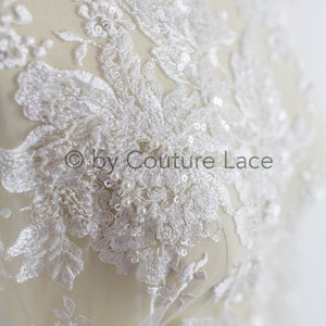 May include: White lace fabric with floral embroidery and pearl and sequin embellishments. The fabric is on a white background. "&copy; by Couture Lace" is printed on the fabric.