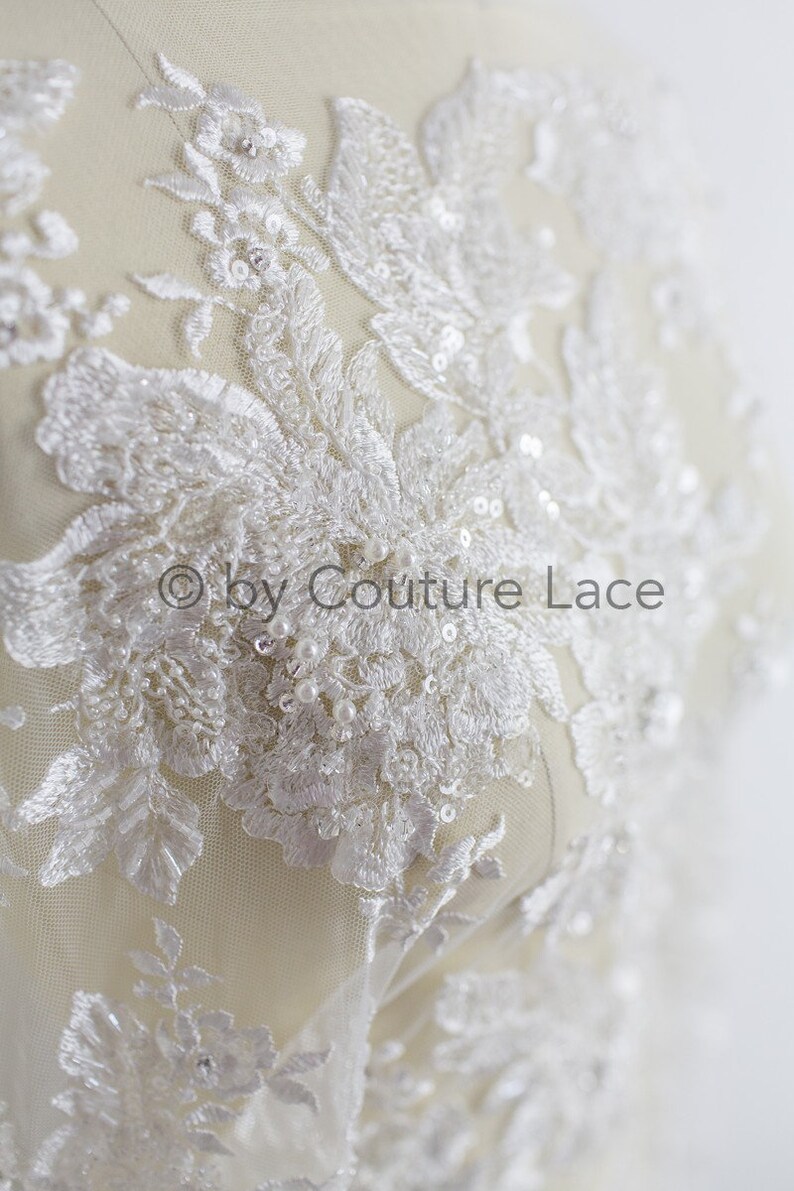 May include: White lace fabric with floral embroidery and pearl and sequin embellishments. The fabric is on a white background. "&copy; by Couture Lace" is printed on the fabric.