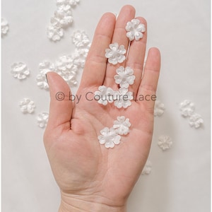 May include: A hand holding a collection of small white fabric flowers. The flowers are scattered on a white surface.