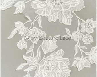 May include: Close-up of white floral lace fabric. The lace features detailed, textured flowers and leaves on a sheer background. The text "© by Couture Lace" is visible.