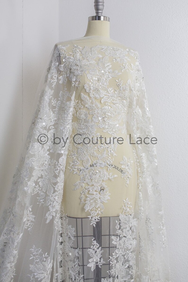 May include: White beaded lace fabric with floral designs and sequins. The fabric is draped over a dress form. The text "by Couture Lace" is visible in the image.