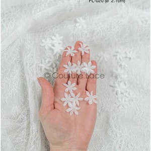 May include: A hand holding a collection of white, delicate, flower-shaped embellishments. The embellishments are approximately 2.1 cm in diameter. FL-020.