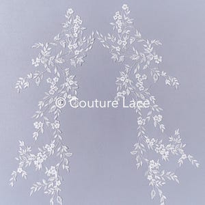 May include: Two white lace appliques with delicate floral designs. Each applique features a vine with small white flowers and leaves. The appliques are perfect for adding a touch of elegance to wedding dresses, bridal accessories, or other craft projects.