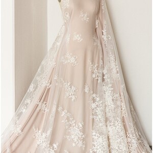 May include: A white lace fabric with a floral pattern draped over a dress form. The lace is sheer and delicate, with a soft, flowing texture. The fabric is a light beige color, and the lace is a bright white.