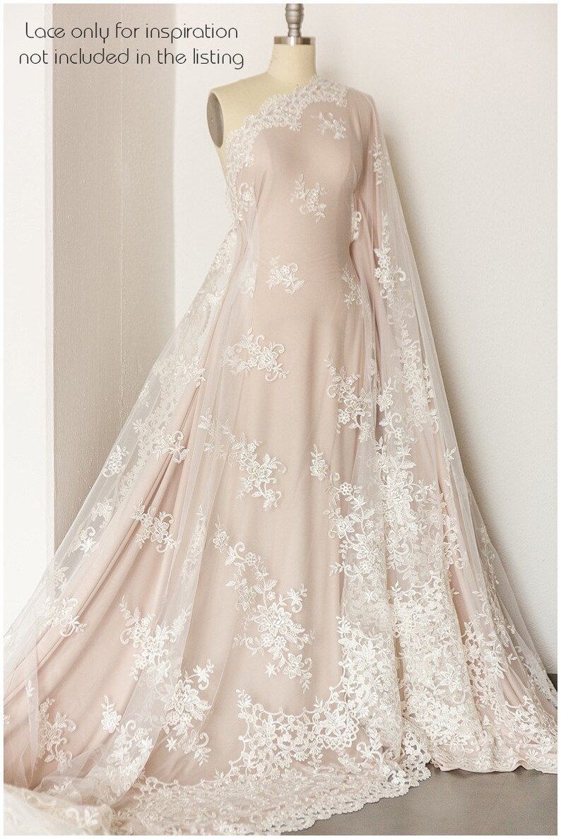 May include: A white lace fabric with a floral pattern draped over a dress form. The lace is sheer and delicate, with a soft, flowing texture. The fabric is a light beige color, and the lace is a bright white.
