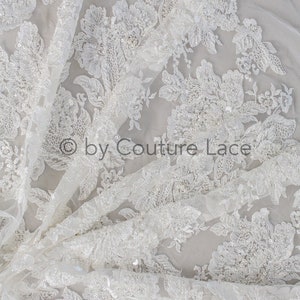 May include: White floral lace fabric with sequins and beads. The lace has a delicate, intricate design and is perfect for wedding dresses, bridal accessories, and other special occasion garments.
