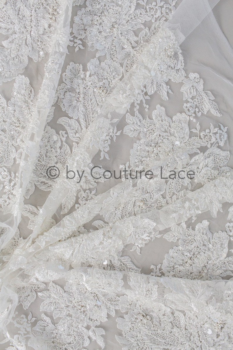 May include: White floral lace fabric with sequins and beads. The lace has a delicate, intricate design and is perfect for wedding dresses, bridal accessories, and other special occasion garments.