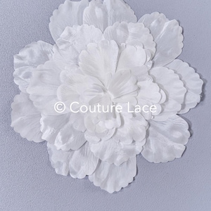 Può includere: Un grande fiore bianco a strati in tessuto, con il testo "© Couture Lace" al centro. Il fiore è posto su uno sfondo grigio chiaro, creando un'estetica morbida ed elegante. I petali sono dettagliati e strutturati.