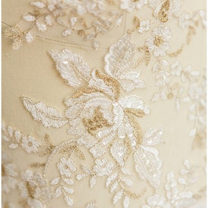 May include: Close-up of a delicate white and gold lace fabric with a floral pattern. The fabric is soft and intricate, with small beads and sequins adding sparkle.