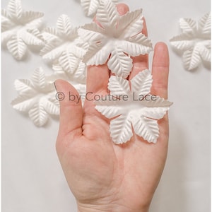 May include: A hand holding several white, paper flower petals. The petals have a ruffled edge and are arranged in a star shape. The text "© by Couture Lace" is visible in the image.