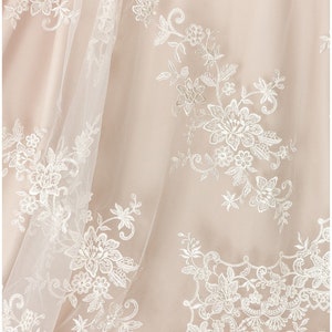 May include: Close-up of a delicate, white lace overlay on a blush-colored fabric. The lace features intricate floral patterns and delicate stitching.
