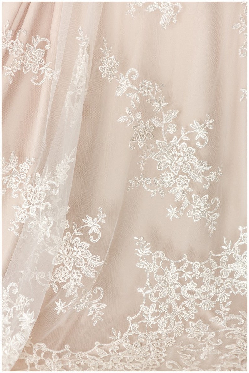 May include: Close-up of a delicate, white lace overlay on a blush-colored fabric. The lace features intricate floral patterns and delicate stitching.