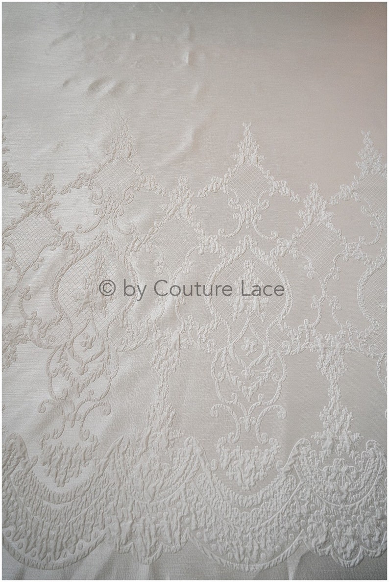 May include: White lace fabric with a delicate, embossed floral pattern. The lace has a scalloped edge and is perfect for adding a touch of elegance to any project.
