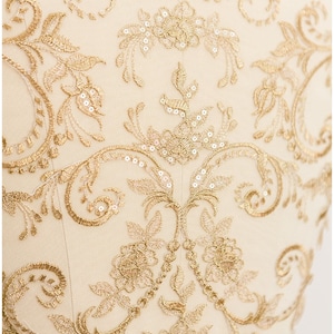 May include: Close-up of cream fabric with detailed gold embroidery. The design includes floral and scroll patterns, with small sequins adding sparkle. The fabric is likely for formal wear or decorative use.