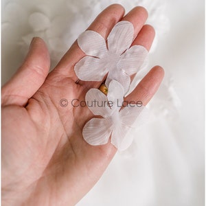 May include: Two white fabric flower appliques, each with five petals, held in a hand against a white background. The text "Couture Lace" is visible in the image.
