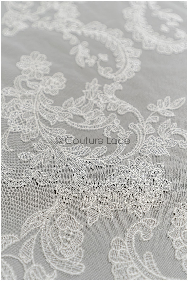 May include: White floral lace fabric with a delicate pattern. The lace is on a sheer mesh background.