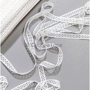 Peut inclure: Galon de dentelle blanche avec un motif de cercle répétitif. Le galon est plié et disposé sur une surface grise.