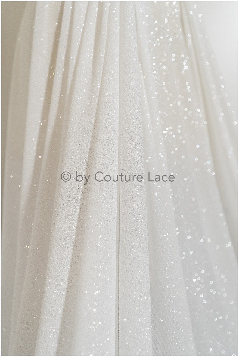 May include: White tulle fabric with a subtle sparkle. The fabric is folded and draped, creating a soft, flowing look. &copy; by Couture Lace