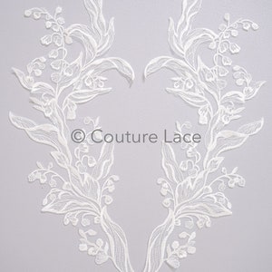 以下が含まれることがあります： ハート型の白いレースのアップリケ2つ。繊細な花柄で、スズランの花と葉がデザインされています。アップリケは、ウェディングドレス、ブライダルアクセサリー、その他のクラフトプロジェクトにエレガントなアクセントを加えるのに最適です。 © Couture Lace