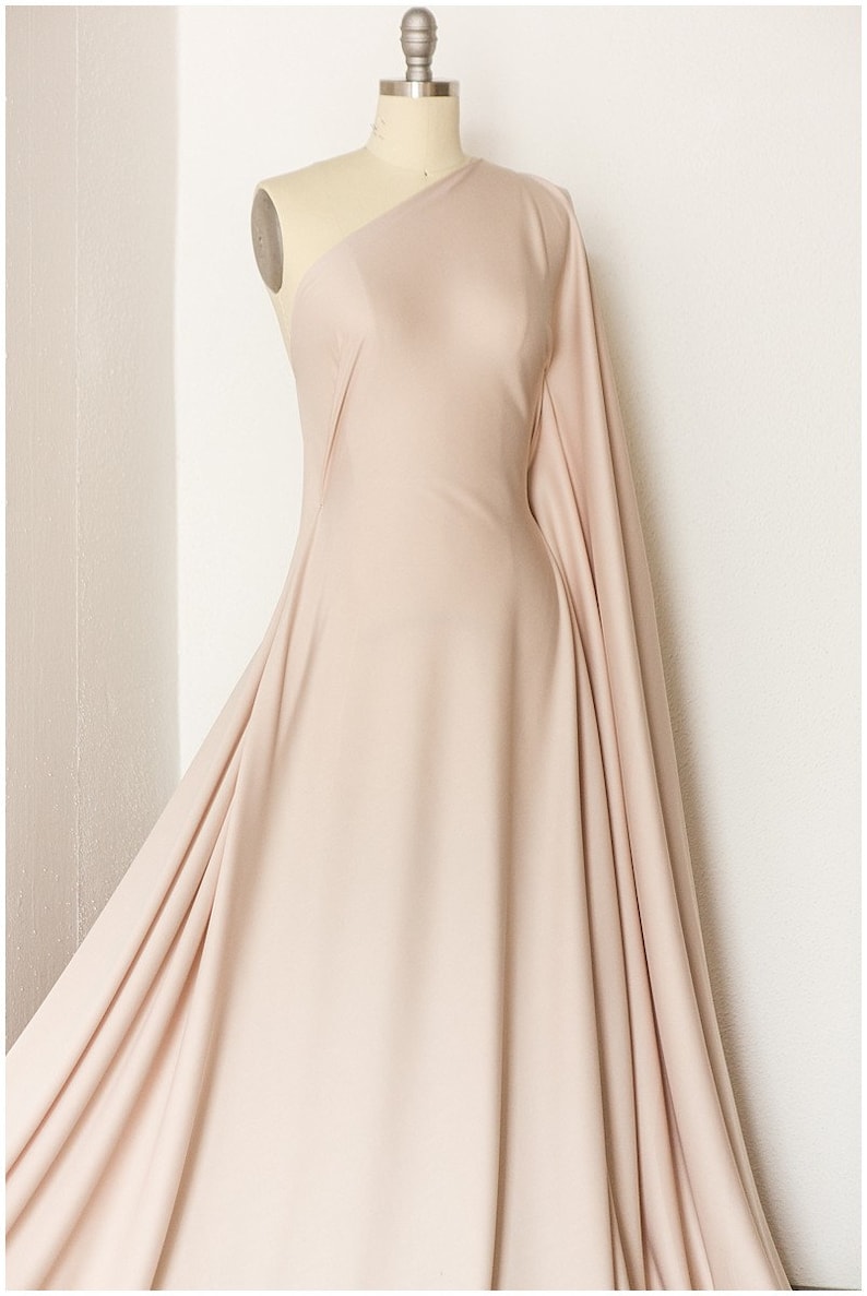 May include: A pale pink fabric draped over a dress form. The fabric is smooth and has a slight sheen. The fabric is gathered at the shoulders and flows down to the floor.