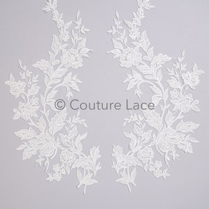 May include: Two white floral lace appliques with intricate designs of flowers and leaves. The appliques are symmetrical and feature the text "Couture Lace". The lace is set against a light gray background.
