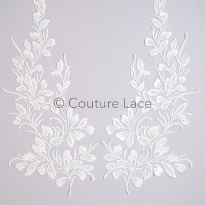May include: Two white lace appliques with a floral design. The appliques are shaped like a vine with leaves and flowers. The appliques are on a light gray background.