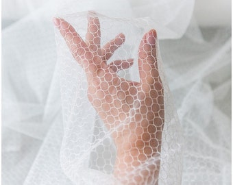 May include: A hand reaches through a white, sheer fabric with a circular pattern. The fabric is delicate and translucent, creating a soft and ethereal effect.