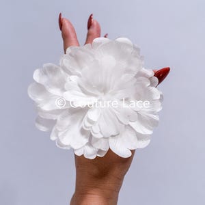 Op de afbeelding: Een grote witte bloem van stof met een gelaagd bloemblaadjesontwerp. De bloem wordt in een hand met rode nagellak vastgehouden.