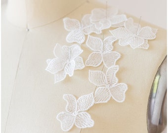 May include: Close-up of a cream-colored dress form with delicate white floral lace appliqués pinned to the shoulder. The lace features detailed petal patterns and is secured with silver pins, showcasing a bridal or formal wear design.