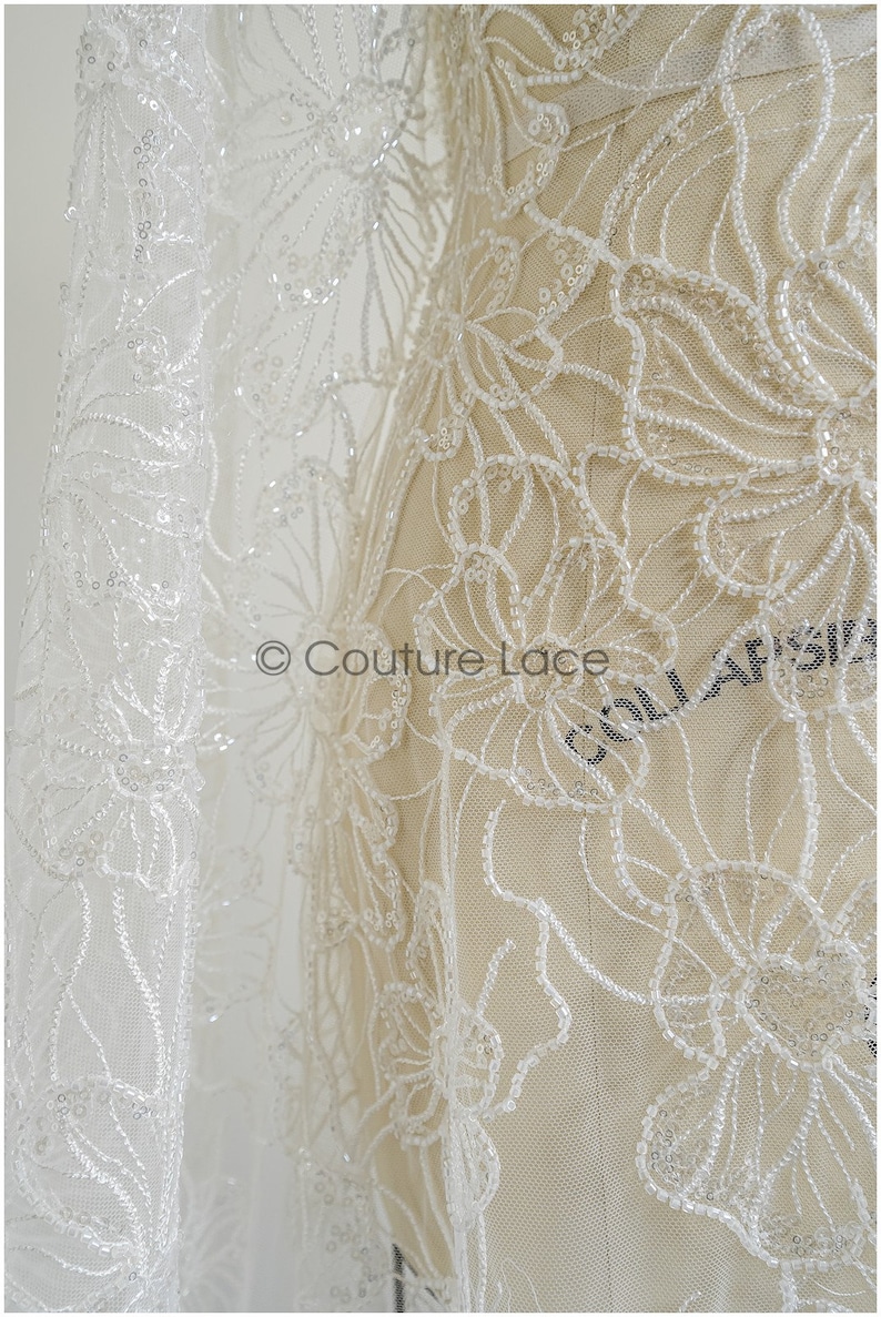 Pu&ograve; includere: Tessuto di pizzo bianco perlato con motivo floreale. Il pizzo &egrave; delicato e intricato, con piccole perline sparse su tutto il tessuto. Il tessuto &egrave; mostrato su un manichino, e il testo "Couture Lace" &egrave; visibile nell'angolo inferiore sinistro.