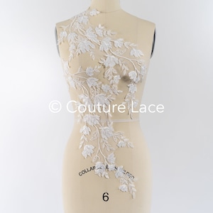 May include: White floral lace applique with delicate leaves and flowers. The lace is displayed on a dress form.