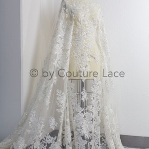May include: White floral lace fabric with sequins and beads. The lace is on a dress form and has a flowing design.  &copy; by Couture Lace