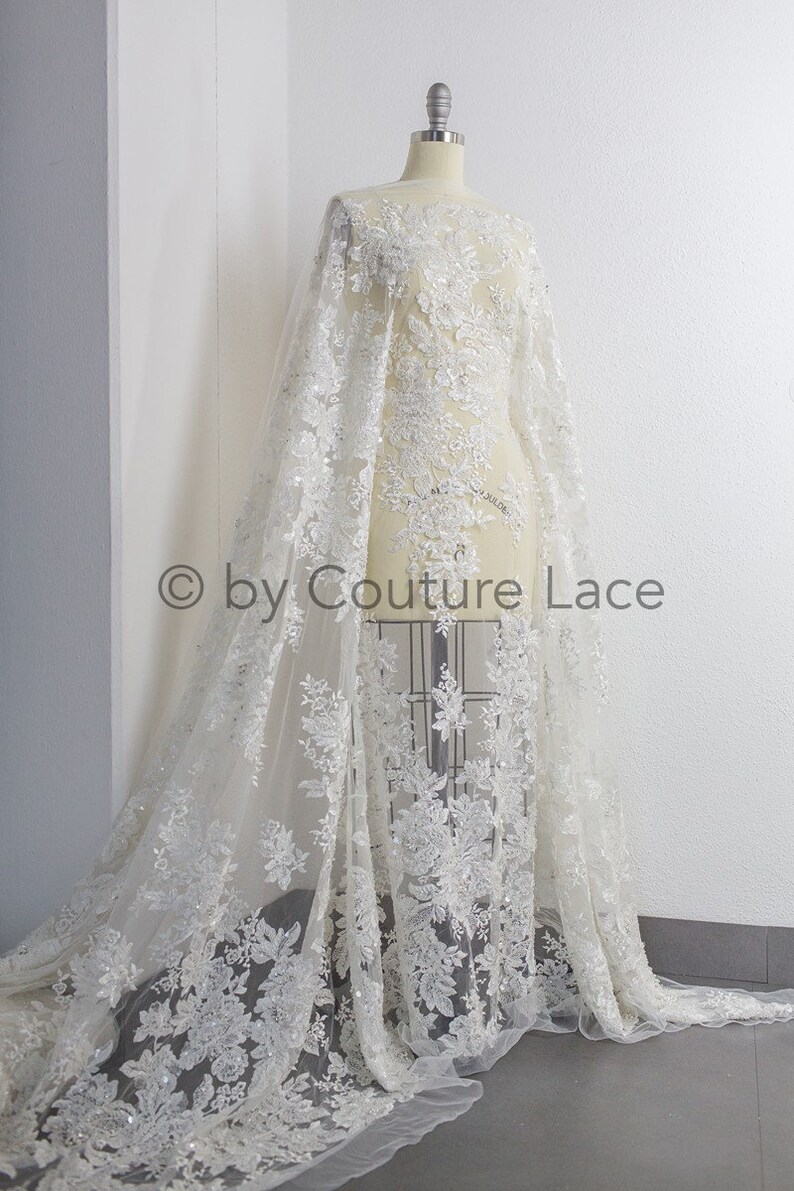 May include: White floral lace fabric with sequins and beads. The lace is on a dress form and has a flowing design.  &copy; by Couture Lace