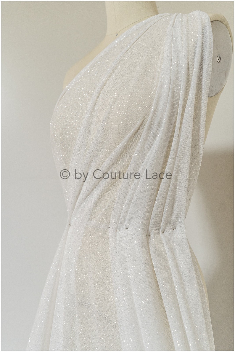 May include: White sheer fabric with a sparkly finish. The fabric is draped over a dress form and has a slight sheen. The fabric is lightweight and has a soft texture. &copy; by Couture Lace