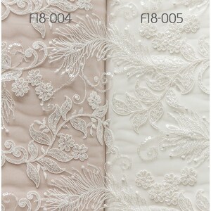 May include: Two samples of white lace fabric with a floral and feather design. The fabric on the left is a light beige color and the fabric on the right is white. The fabric on the left is labeled "F18-004" and the fabric on the right is labeled "F18-005".