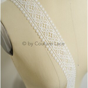 Puede incluir: Borde de encaje blanco con borde festoneado y un delicado patr&oacute;n floral.  "&copy; by Couture Lace" est&aacute; impreso en el borde.