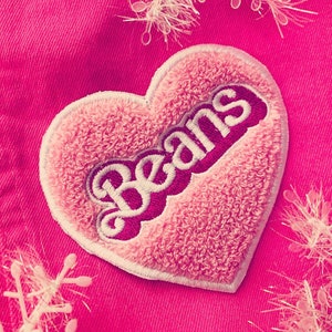 Pode incluir: Patch rosa em forma de coração com costuras brancas e a palavra "Beans" em letras brancas sobre um fundo rosa.