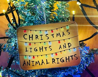 Puede incluir: Adorno navideño rectangular con el texto "CHRISTMAS LIGHTS AND ANIMAL RIGHTS" en letras mayúsculas negras. El adorno tiene una hilera de luces de colores en la parte superior. Cuelga de un árbol de Navidad de oropel azul.