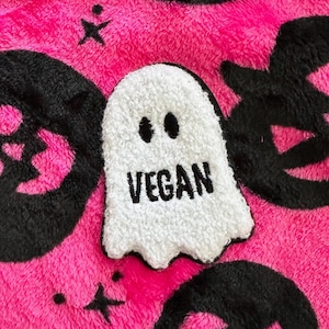 Op de afbeelding: Een witte pluche geest met zwarte ogen en het woord "VEGAN" erop geborduurd. De geest bevindt zich op een roze pluche achtergrond met zwarte schedelpatronen.