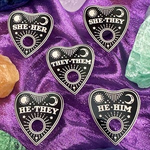 May include: Five black heart-shaped pins with white celestial designs and text. Each pin features a crescent moon, sunburst, and text indicating pronouns: "SHE HER", "SHE THEY", "THEY THEM", "HE HIM", and "HE THEY".