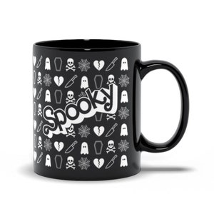 May include: Black ceramic mug with a white "Spooky" design. The mug features a repeating pattern of Halloween-themed icons, including ghosts, spiderwebs, skulls, knives, and coffins. The mug has a curved handle and a glossy finish.