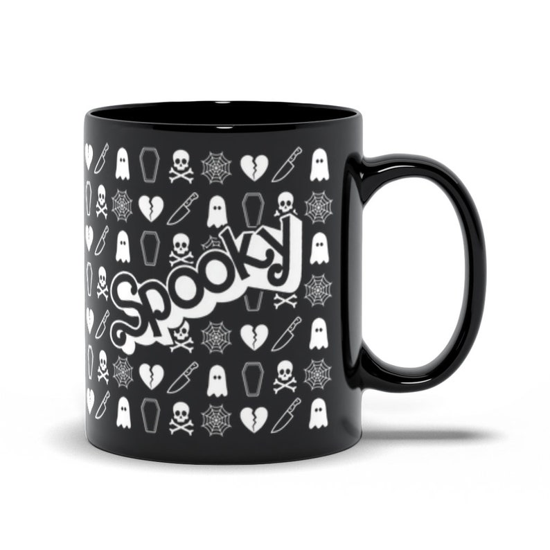 May include: Black ceramic mug with a white "Spooky" design. The mug features a repeating pattern of Halloween-themed icons, including ghosts, spiderwebs, skulls, knives, and coffins. The mug has a curved handle and a glossy finish.
