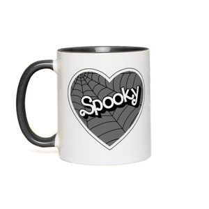 May include: White coffee mug with a black rim and handle. The mug features a black and white graphic of a heart with a spiderweb design and the word "Spooky" in a retro font.