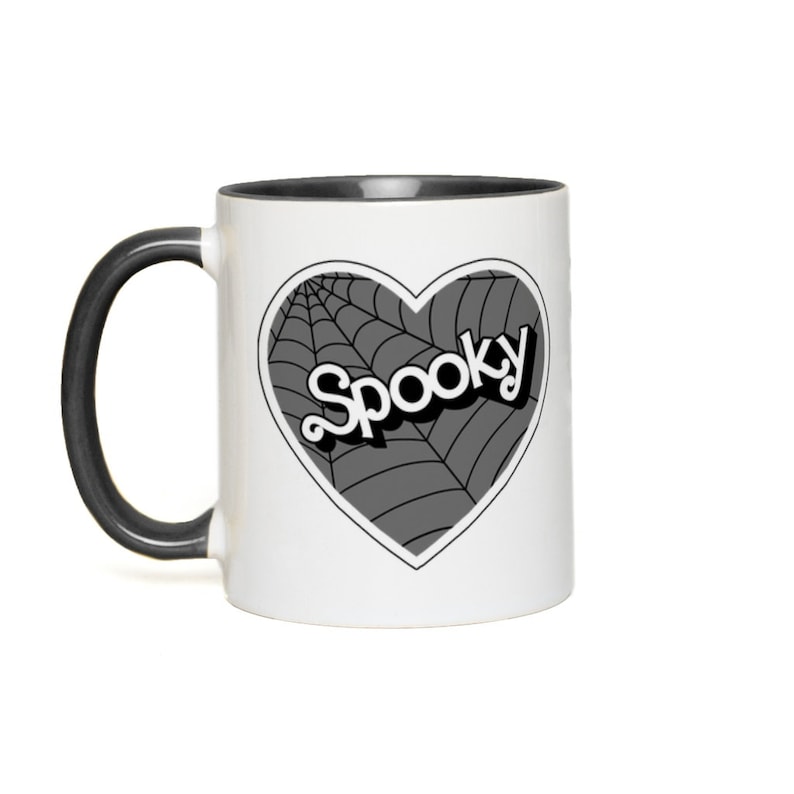 May include: White coffee mug with a black rim and handle. The mug features a black and white graphic of a heart with a spiderweb design and the word "Spooky" in a retro font.