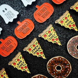 May include: A collection of enamel pins featuring various food items like donuts, pizza slices, and pumpkins. Each pin has a different text on it, including "They/Them Vegan", "She/They Vegan", and "He/Him".
