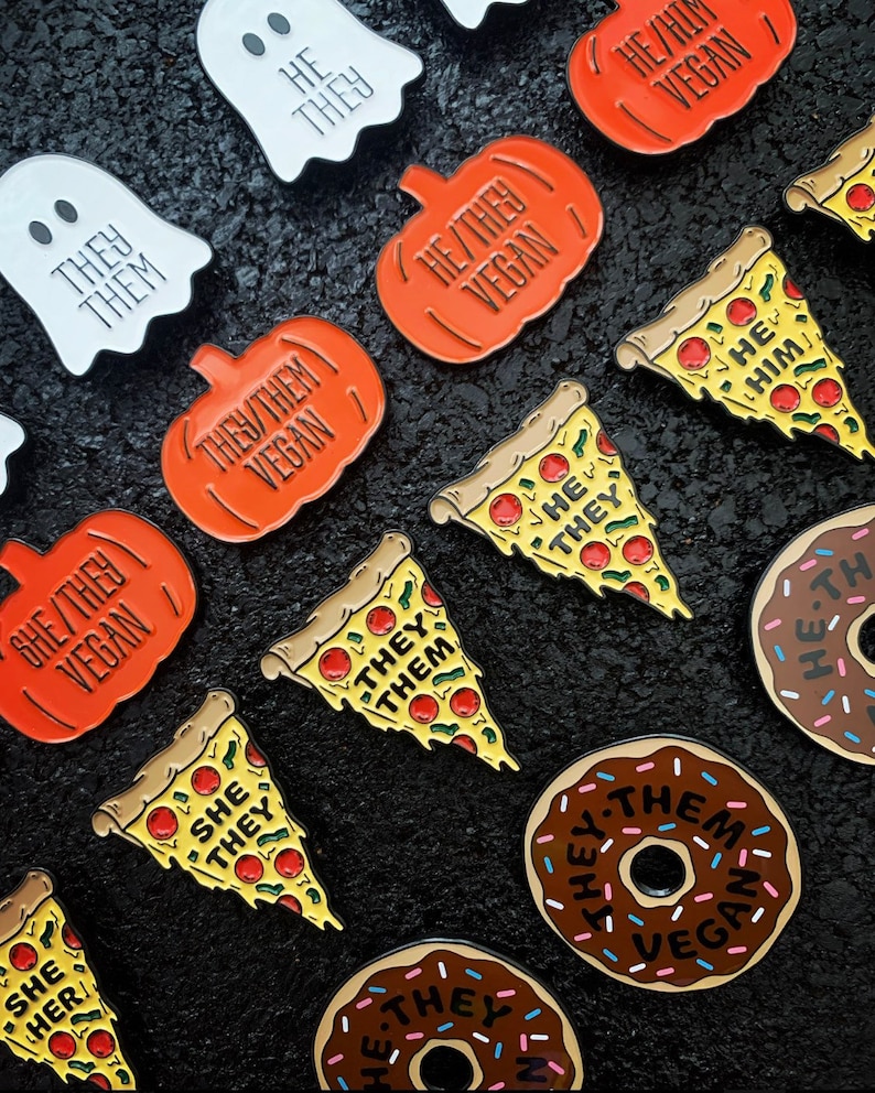 May include: A collection of enamel pins featuring various food items like donuts, pizza slices, and pumpkins. Each pin has a different text on it, including "They/Them Vegan", "She/They Vegan", and "He/Him".